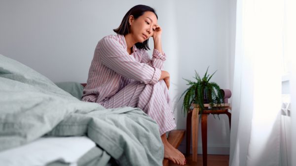 ベッドの上で膝をかかえ、頬杖をついて悩む女性